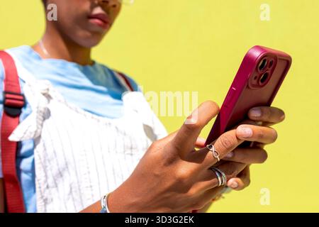Une image en gros plan d'une femme noire utilisant un smartphone sur un fond jaune vif. Elle est concentrée sur son appareil, portant une tenue décontractée et déco Banque D'Images
