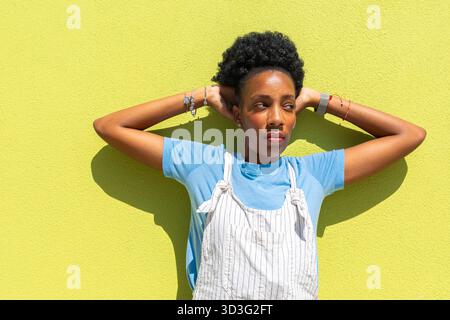 Une femme noire se tient contre un mur jaune vif, portant une chemise bleue et une salopette rayée. Elle place désinvolte ses mains derrière sa tête, trempant Banque D'Images