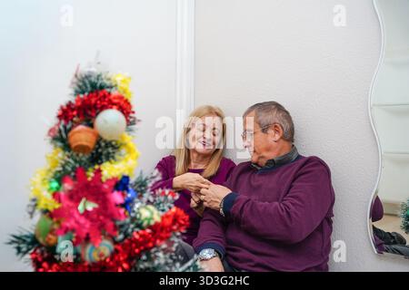 Un couple d'âge mûr partage un moment tendre à côté d'un arbre de Noël décoré de couleurs vives, célébrant la tradition et la convivialité dans un cadre confortable durin Banque D'Images