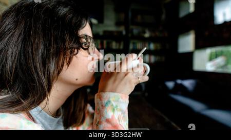 Une femme portant des lunettes profite d'une tasse chaude de chocolat chaud dans un cadre confortable. L'éclairage tamisé de la pièce et les livres qui l'entourent créent une atmosphère relaxante Banque D'Images