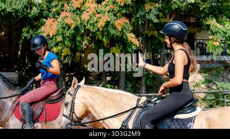 Deux filles portant des casques montent à cheval pendant l'entraînement tout en utilisant leur smartphone. Un mélange de technologie et de style de vie équestre, capturé sur un ensoleillé Banque D'Images