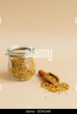 Granulés de pollen d'abeille biologiques dans un bocal en verre rustique accompagnés d'une pelle en bois sur un fond beige, mettant en valeur un opti de superaliment naturel et sain Banque D'Images