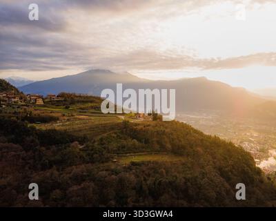 Vue aérienne d'un coucher de soleil doré illuminant les collines ondulantes et un petit village perché au sommet, projetant des ombres sur la vallée en contrebas, Tenno, Trentin Banque D'Images