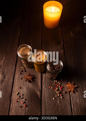 Une nature morte confortable de pots d'épices en verre, de grains de poivre colorés et d'anis étoilé disposés sur une surface en bois rustique, doucement éclairés par une bougie incandescente Banque D'Images