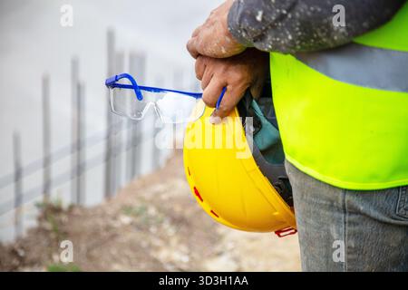 Travailleur tenant un casque jaune et des lunettes de sécurité sur le chantier de construction à l'extérieur, portant un gilet de sécurité. Banque D'Images
