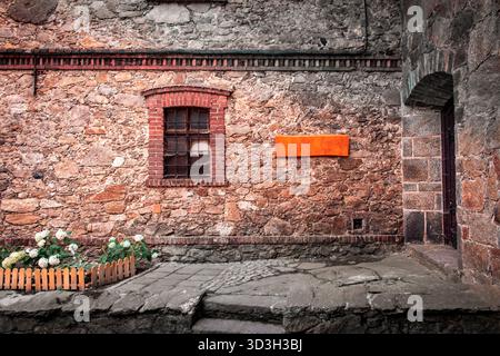 Un vieux mur de pierre du château de Czocha avec une petite fenêtre, un panneau orange vif, un parterre de fleurs clôturé avec des fleurs blanches, et un chemin de pierre. Banque D'Images