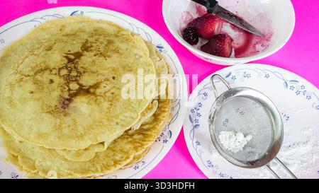 Une pile de crêpes maison se trouve sur une assiette à côté d'un bol de fraises fraîches et d'un tamis avec du sucre en poudre. Un couteau repose à côté du bol, lire Banque D'Images