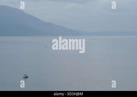 Petit bateau flottant sur un lac calme et vaste avec des montagnes brumeuses en arrière-plan. Banque D'Images