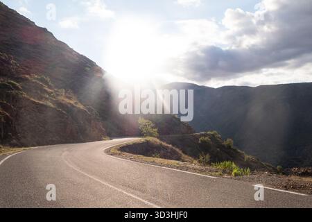 Route de montagne sinueuse qui traverse un paysage rocheux spectaculaire avec la lumière du soleil qui traverse les nuages au-dessus des vallées profondes. Banque D'Images