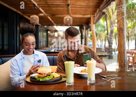 Un couple diversifié sur une date gourmande aime manger décontracté et un moment de téléphone, partageant des vibrations ensoleillées, des boissons Banque D'Images