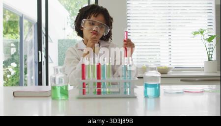 Enfant en blouse de laboratoire examinant un tube à essai rouge sur le comptoir de cuisine, avec un portoir pour tubes à essai Banque D'Images
