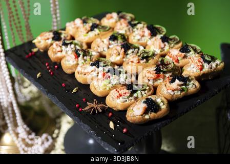 Nourriture, canapés de fruits de mer élégants surmontés de crevettes, caviar, herbes et mélange de salade, magnifiquement disposés sur un plateau de service sombre Banque D'Images