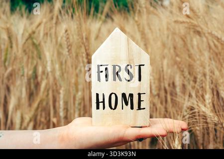 Main tenant un panneau en bois qui dit First Home. Le signe est placé dans un champ de hautes herbes Banque D'Images