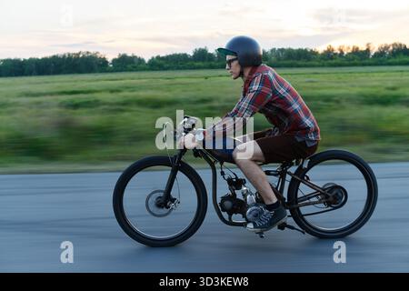 Portrait vue de côté d'un jeune homme hipster chevauchant un vélo rétro sur une route de campagne déserte au crépuscule Banque D'Images