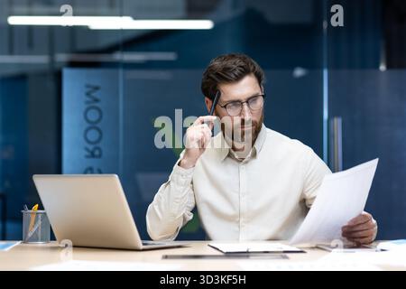 Un jeune homme sérieux et concentré est assis à un bureau dans le bureau et étudie les documents qu'il tient dans sa main. Banque D'Images