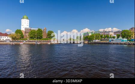 Vyborg, Russie - 27 mai 2024 : une scène côtière pittoresque avec un château blanc avec un dôme vert, des murs historiques, des arbres luxuriants et des bâtiments colorés Banque D'Images