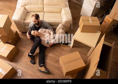 Couple paisible se détend sur le sol près du canapé avec des boîtes en carton emballées autour d'eux. La vue aérienne montre les conjoints jouissant d'une communication calme et de liens pendant le jour du déménagement dans le nouvel appartement. Banque D'Images