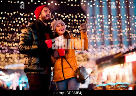Joyeux couple sous les lumières festives profite de boissons chaudes au marché de nuit célébrant la magie de noël ensemble Banque D'Images