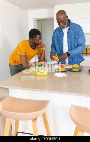 Père afro-américain et fils assemblant des hamburgers à l'îlot de cuisine, avec planche à découper Banque D'Images
