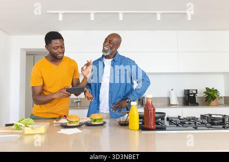 Cuisine afro-américaine père et fils préparant des hamburgers dans la cuisine moderne, à l'aide d'un appareil de tablette Banque D'Images