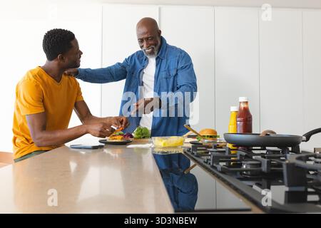 Construire père et fils afro-américains préparant des hamburgers dans la cuisine, avec smartphone et casserole Banque D'Images