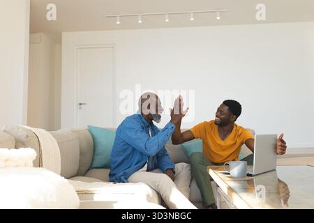 Père et fils afro-américains chevronnés assis sur le canapé dans le salon, avec ordinateur portable et tasse Banque D'Images