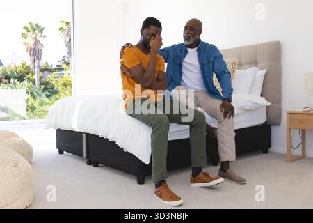 Père afro-américain réconfortant fils assis sur le lit dans une chambre moderne, avec des draps blancs Banque D'Images