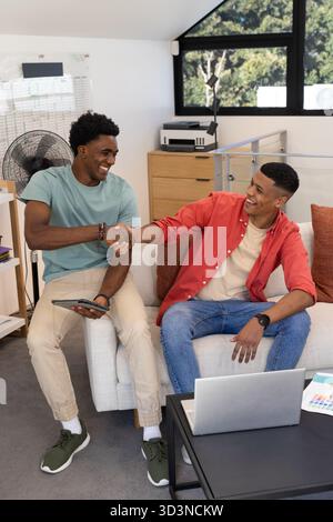 Serrant la main divers collègues masculins assis sur un canapé blanc dans le bureau à domicile, avec tablette et ordinateur portable Banque D'Images