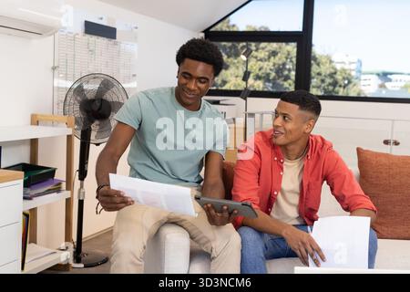 Divers amis masculins examinant les feuilles imprimées et utilisant la tablette sur le canapé loft, avec tableau blanc Banque D'Images
