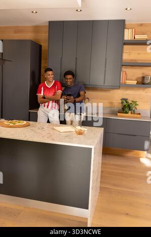 Pencher divers amis masculins souriant et regardant smartphone à l'îlot de cuisine, avec pizza Banque D'Images