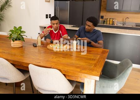 Divers amis masculins mangeant des tranches de pizza sur une planche à découper à table dans la cuisine, avec des boissons Banque D'Images