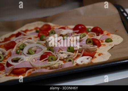 Pizza maison préparée sur une plaque de cuisson avec légumes frais, jambon, champignons, tomates, oignons, et des olives. Cette image est parfaite pour les blogs de cuisine, Banque D'Images