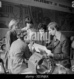 Une photo d'archive de 1981 montre une réunion de production à l'AIZ (armature-Insulator Plant) à Sloviansk, RSS d'Ukraine. Un ingénieur ou contremaître masculin détient un dessin technique et instruit une équipe de quatre travailleuses. Ils sont rassemblés autour d'un grand composant industriel, éventuellement le stator d'un moteur électrique. La scène se déroule dans l'atelier, directement devant une grande murale emblématique de propagande soviétique représentant l'industrie, la science (atomes) et le progrès. Cela capture le travail industriel et la culture visuelle idéologique dans le Donbass pacifique avant la guerre Banque D'Images
