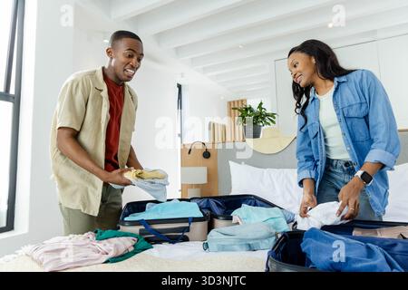 Couple afro-américain emballant et pliant des vêtements dans la chambre, avec valises ouvertes et sac à dos Banque D'Images
