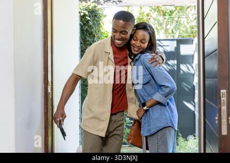 Couple afro-américain debout serrant à la porte d'entrée ouverte, avec valise, sac à main et porte-clés Banque D'Images