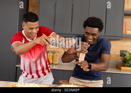 Moudre divers amis masculins assaisonnant la pâte à pizza à l'îlot de cuisine, avec des moulins à épices Banque D'Images