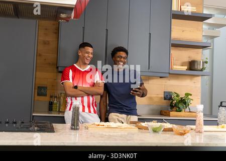 Penchant divers amis masculins vérifiant smartphone à l'île dans la cuisine moderne, avec de la pâte à pizza Banque D'Images