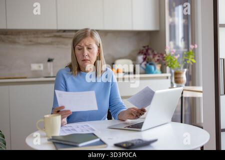 Femme senior à la table de cuisine à la maison se sentant stressée et confus tout en regardant plusieurs factures et documents financiers, travaillant sur son ordinateur portable pour gérer les finances personnelles Banque D'Images