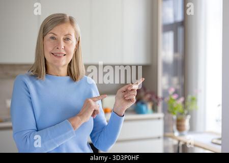 Femme senior souriante et pointant sur le côté dans une cuisine moderne, présentant un espace de copie vide pour la publicité ou l'information, portrait domestique joyeux et détendu Banque D'Images
