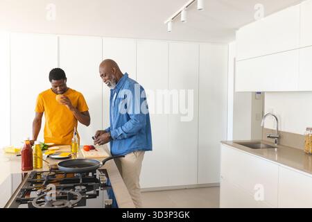 Père afro-américain et fils coupant des légumes dans une cuisine moderne, avec poêle et tasse Banque D'Images