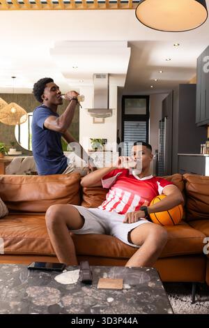 Relaxez divers amis masculins en sirotant des boissons sur le canapé bronzé dans le salon décloisonné, avec basket-ball Banque D'Images