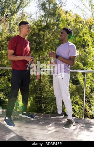 Bavarder divers amis masculins debout sur le pont à l'extérieur, tenant des bouteilles d'eau à la rampe métallique Banque D'Images