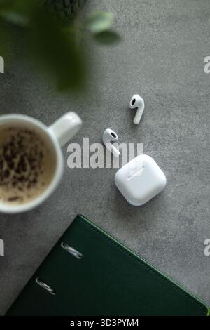 Vue de dessus d'un espace de travail relaxant avec un café mousseux dans une tasse blanche à côté d'écouteurs sans fil blancs et de leur étui, avec un carnet vert sur un gre Banque D'Images