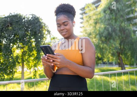 Une femme souriante aux cheveux bouclés profite d'une journée ensoleillée dans le parc, en vérifiant son smartphone, en portant un soutien-gorge de sport et des écouteurs, mettant en vedette un actif et co Banque D'Images