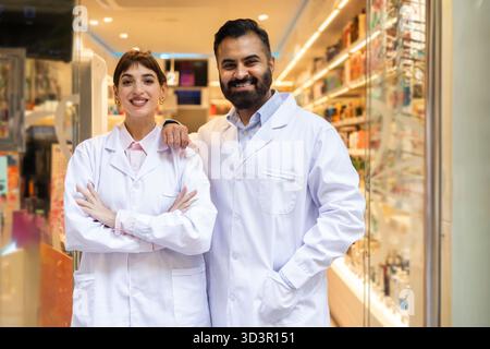 Deux pharmaciens en blouse blanche sourient chaleureusement dans une pharmacie moderne. Ils se tiennent confiants, mettant en valeur une atmosphère accueillante et professionnalisme dans Banque D'Images