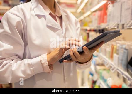 Une pharmacienne portant un manteau blanc utilise un comprimé dans une allée de pharmacie, gérant les stocks et vérifiant les ordonnances. Étagères avec divers soins de santé Banque D'Images
