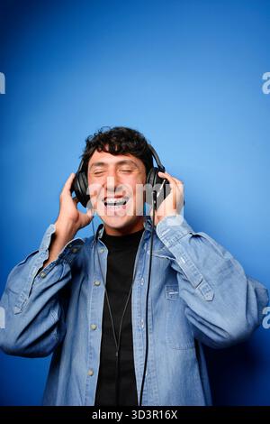 Un jeune homme avec des bretelles porte joyeusement des écouteurs sur un fond bleu, les yeux fermés et souriant largement, véhiculant un sentiment de bonheur et e Banque D'Images