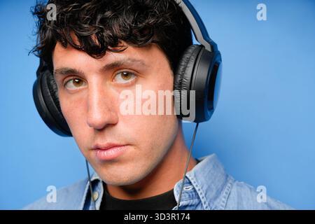 Jeune homme aux cheveux bouclés portant des écouteurs noirs, debout sur un fond bleu vif. Son expression sérieuse et sa chemise en denim décontractée créent un Banque D'Images