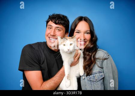 Un jeune couple joyeux pose avec leur adorable chat sur un fond bleu vif. Le trio rayonne de bonheur et de chaleur, capturant l'essence Banque D'Images