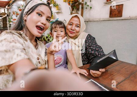 Un moment joyeux de trois générations, une grand-mère musulmane, une mère et une fille d'Asie du Sud-est, capturant un selfie heureux ensemble dans un décor confortable Banque D'Images